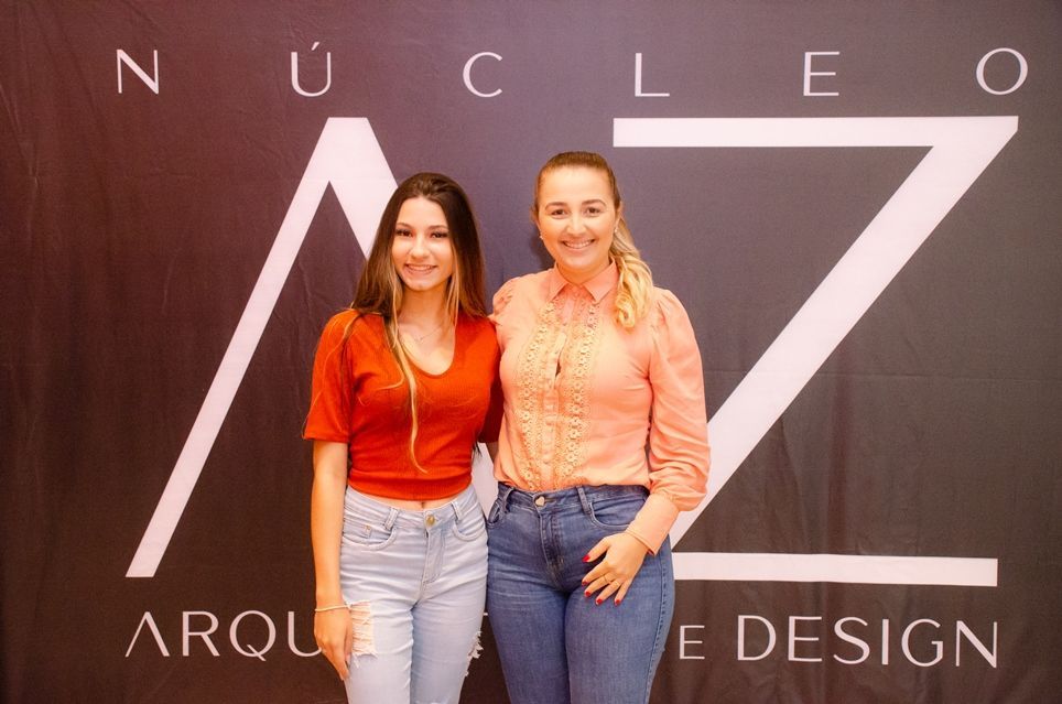 Duas mulheres posam para uma foto em frente a uma placa que diz nucleo arquitetura design