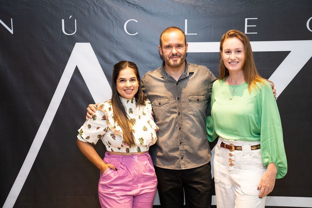 Um homem e duas mulheres estão posando para uma foto em frente a uma placa.