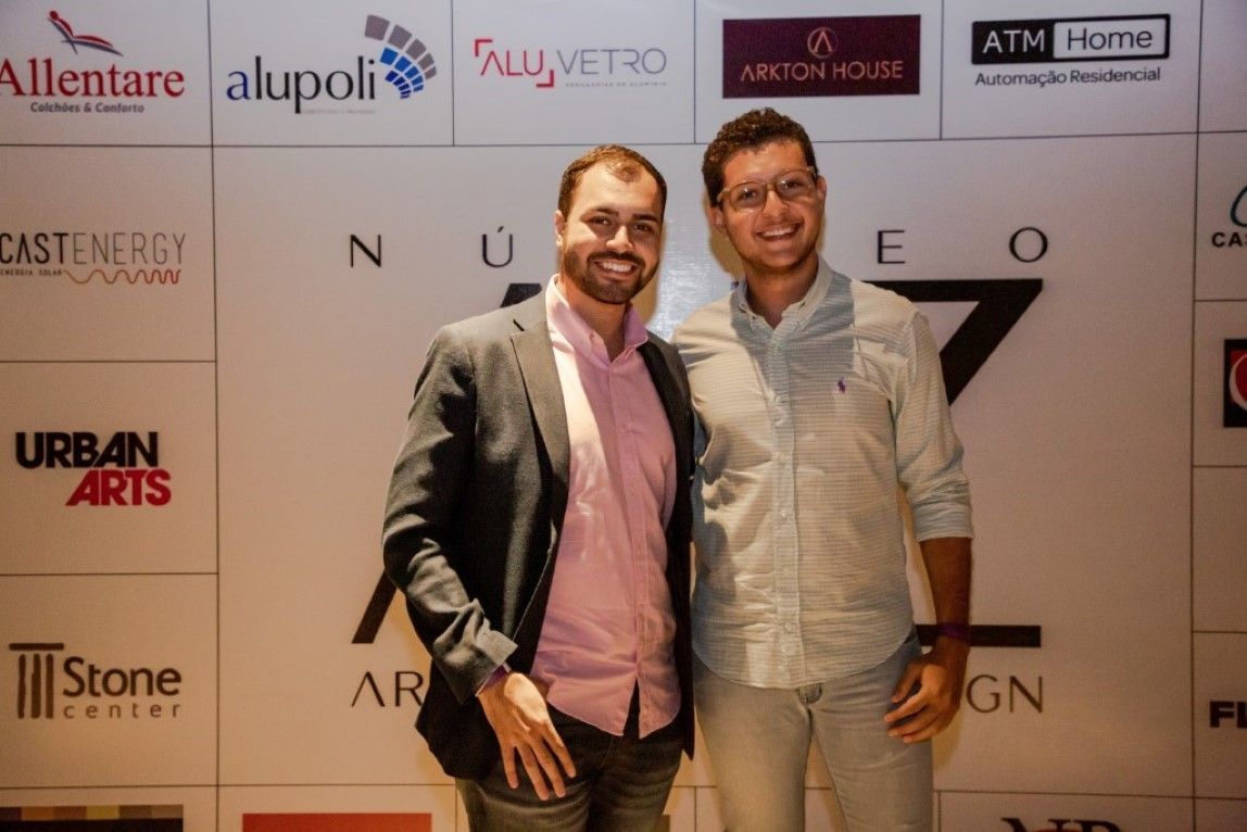 Dois homens posando para uma foto em frente a uma parede com logotipos de diferentes empresas