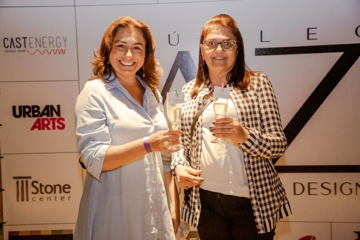 Duas mulheres estão de pé, uma ao lado da outra, segurando taças de vinho.