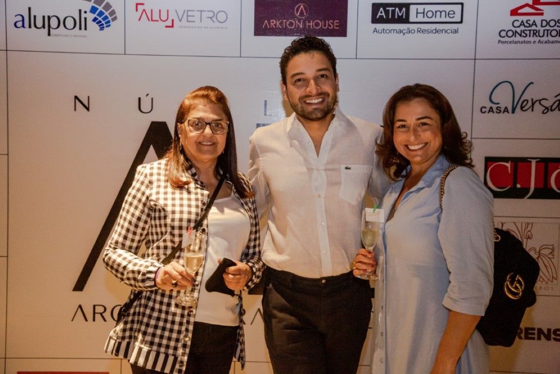 Um homem e duas mulheres posam para uma foto em frente a uma parede com logotipos de várias empresas