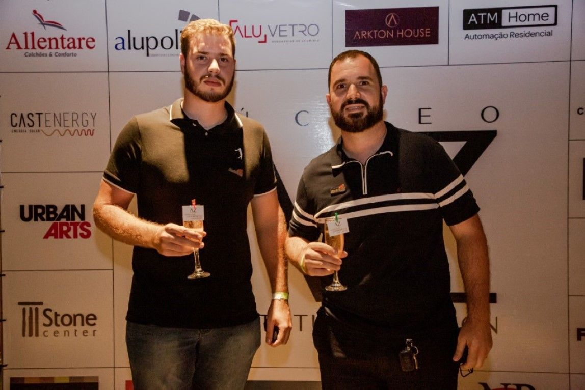 Dois homens estão de pé, um ao lado do outro, segurando taças de vinho em frente a uma parede com logotipos.