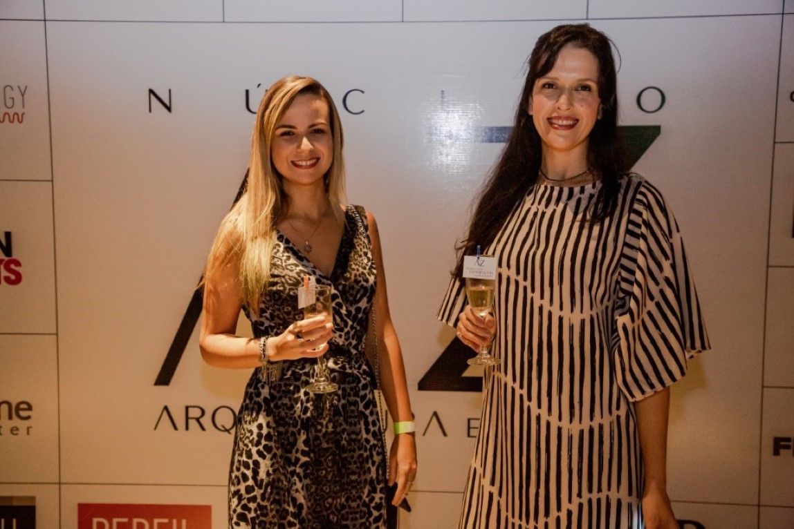 Duas mulheres estão de pé, uma ao lado da outra, em frente a uma parede, segurando taças de champanhe.