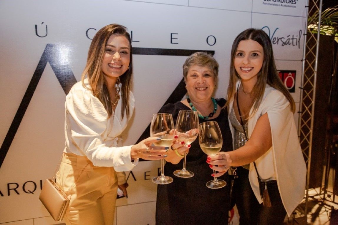 Três mulheres estão brindando com taças de vinho em frente a uma parede.
