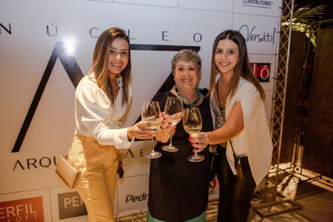 Três mulheres estão de pé, uma ao lado da outra, segurando taças de vinho.