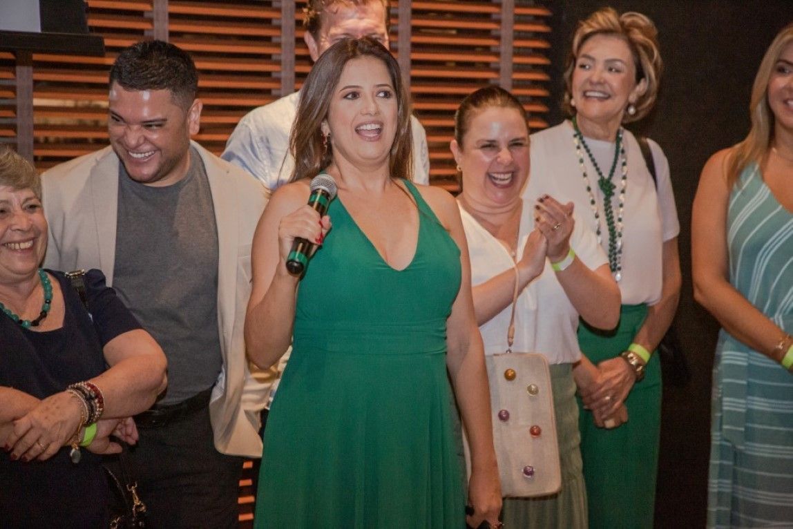 Uma mulher de vestido verde segura um microfone na frente de um grupo de pessoas.