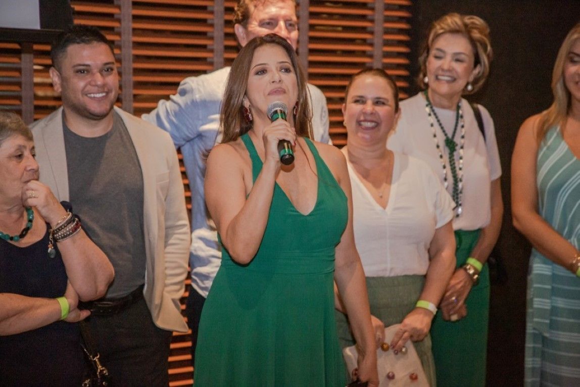 Um grupo de pessoas está em volta de uma mulher de vestido verde segurando um microfone.