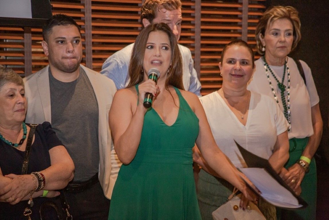 Uma mulher de vestido verde segura um microfone na frente de um grupo de pessoas.