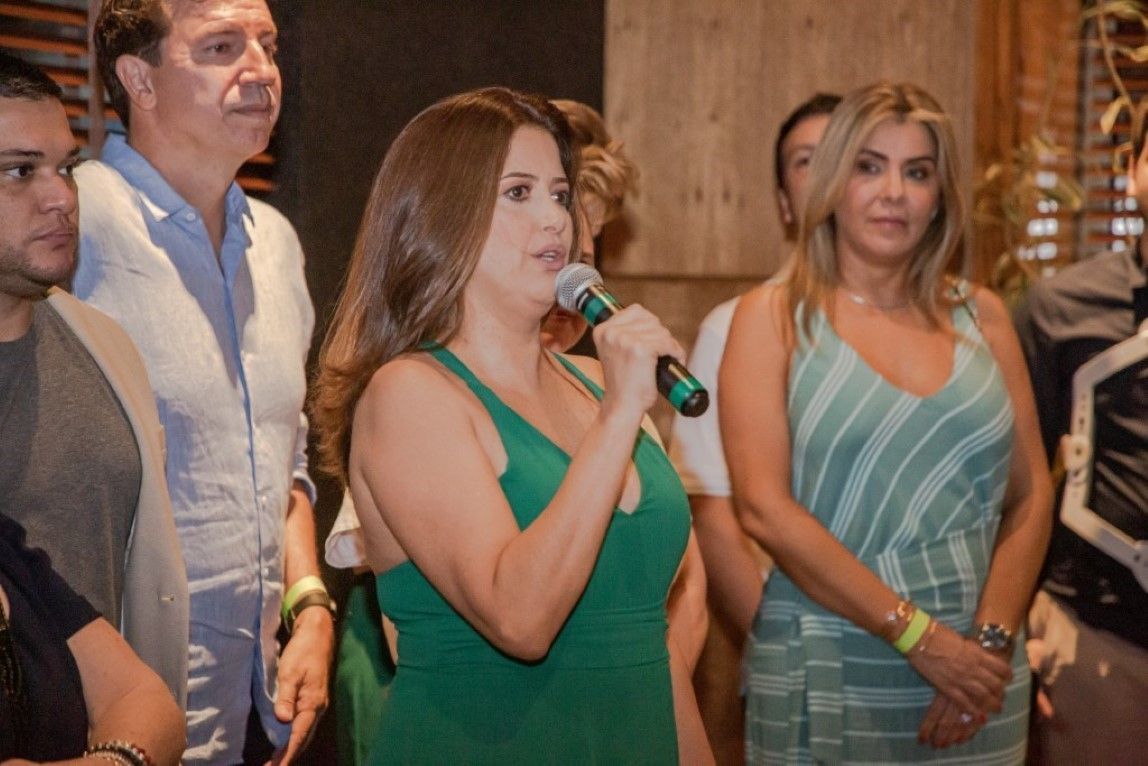Uma mulher de vestido verde segura um microfone na frente de um grupo de pessoas.