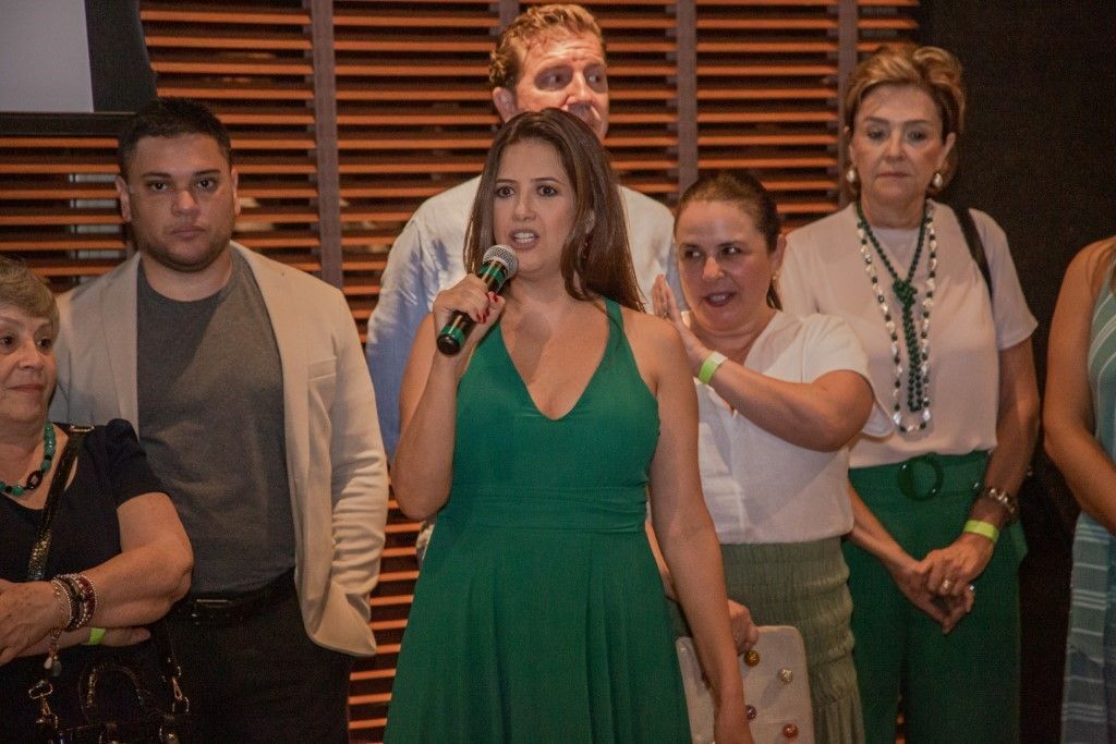 Uma mulher de vestido verde segura um microfone na frente de um grupo de pessoas.