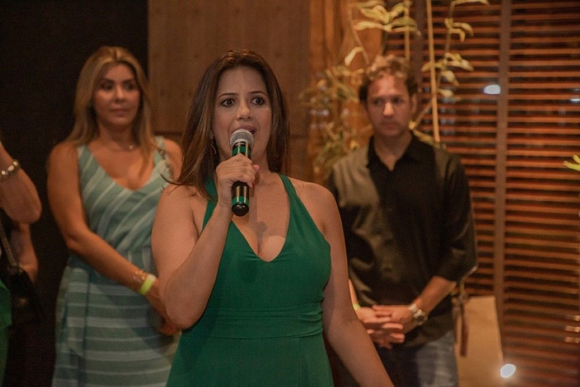 Uma mulher de vestido verde segura um microfone na frente de um grupo de pessoas.