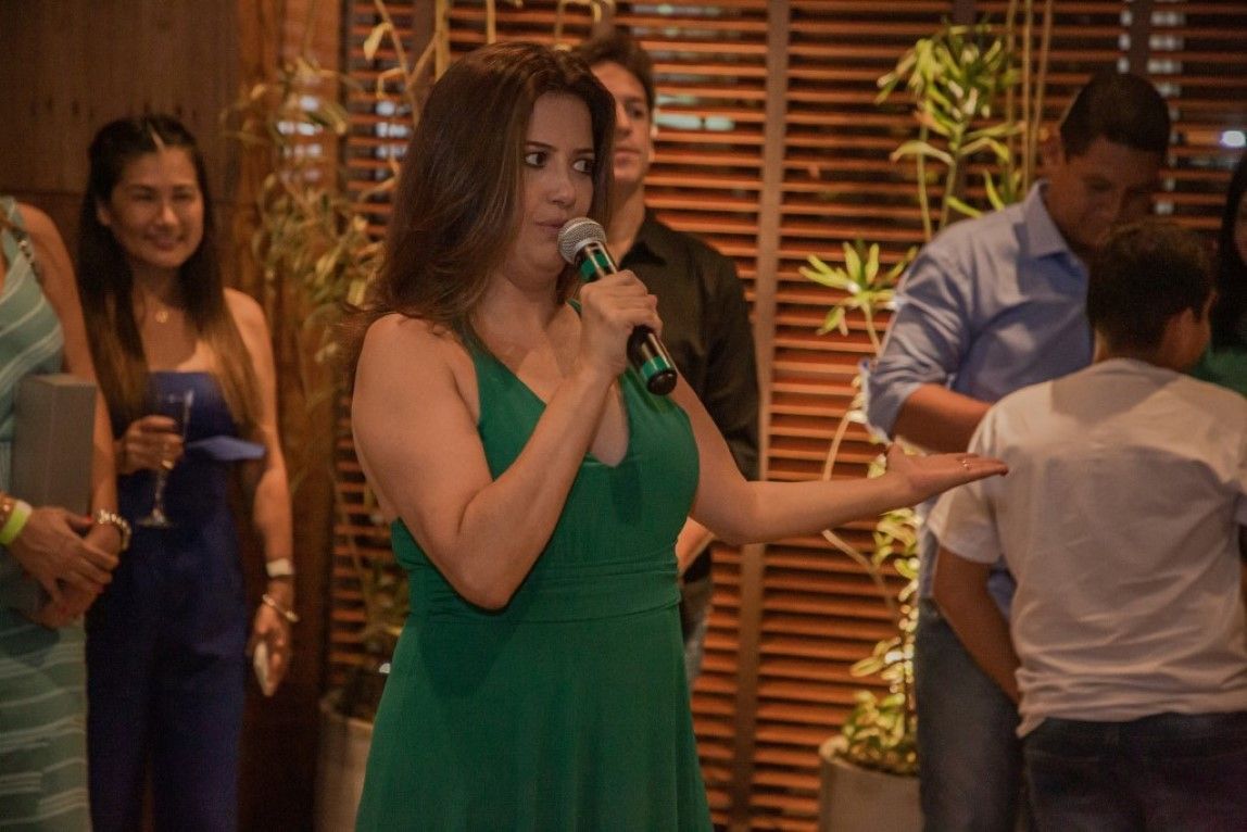 Uma mulher de vestido verde segura um microfone na frente de um grupo de pessoas.
