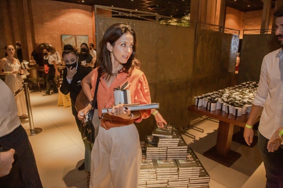 Uma mulher está segurando um livro na mão em frente a uma pilha de livros.