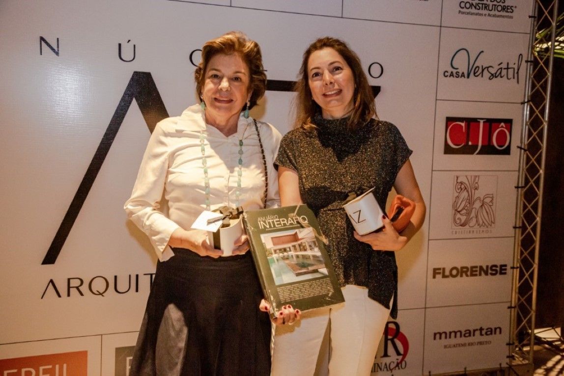 Duas mulheres estão de pé, uma ao lado da outra, segurando livros em frente a uma parede com logotipos.