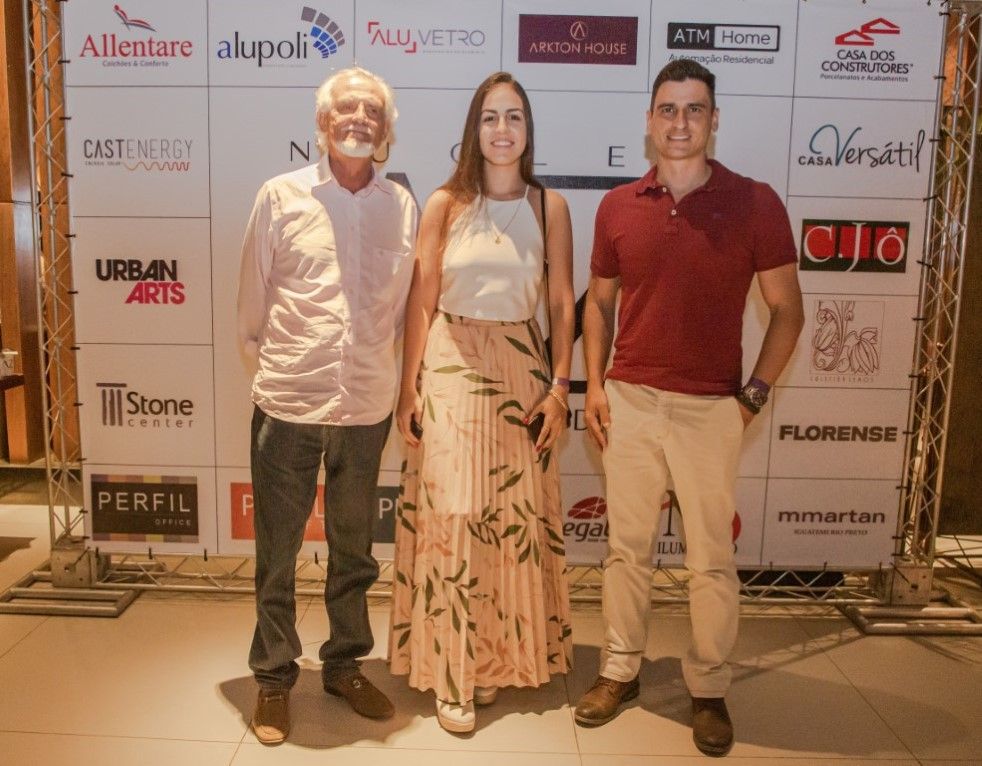 Três pessoas estão posando para uma foto em frente a uma parede com logotipos.