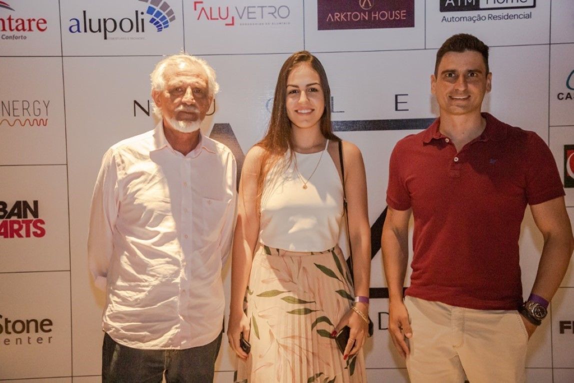 Três pessoas estão posando para uma foto em frente a uma parede com logotipos.