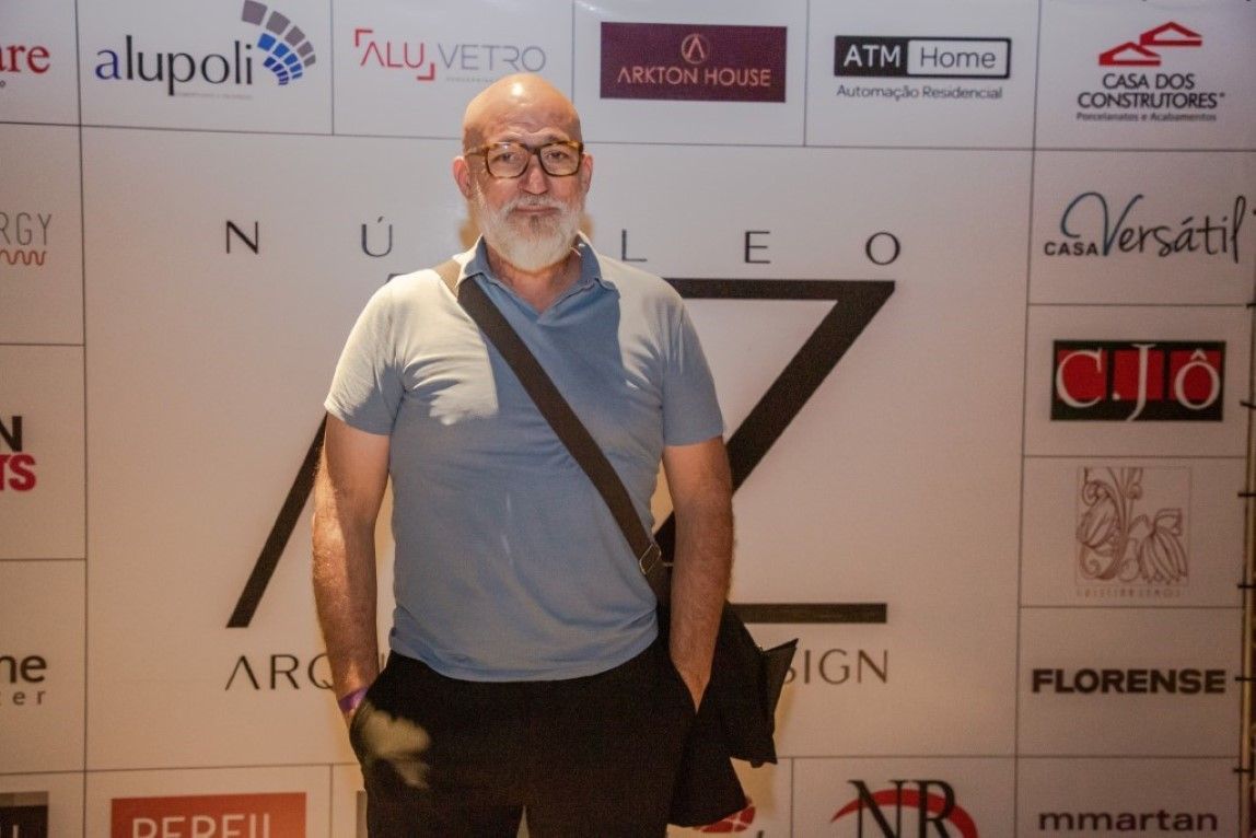 Um homem de óculos e barba está parado em frente a uma parede com logotipos