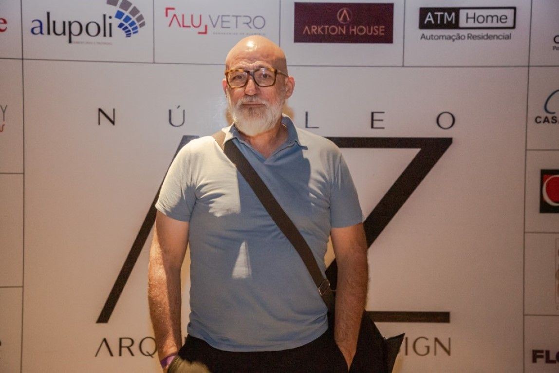Um homem com óculos e barba está em frente a uma parede com logotipos de alupoli e caixa eletrônico