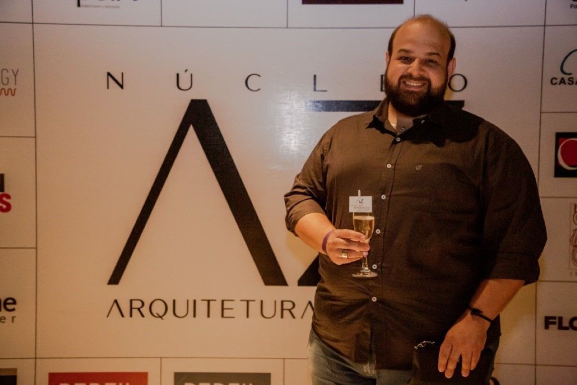 Um homem com barba segura uma taça de champanhe em frente a uma placa que diz arquiteta
