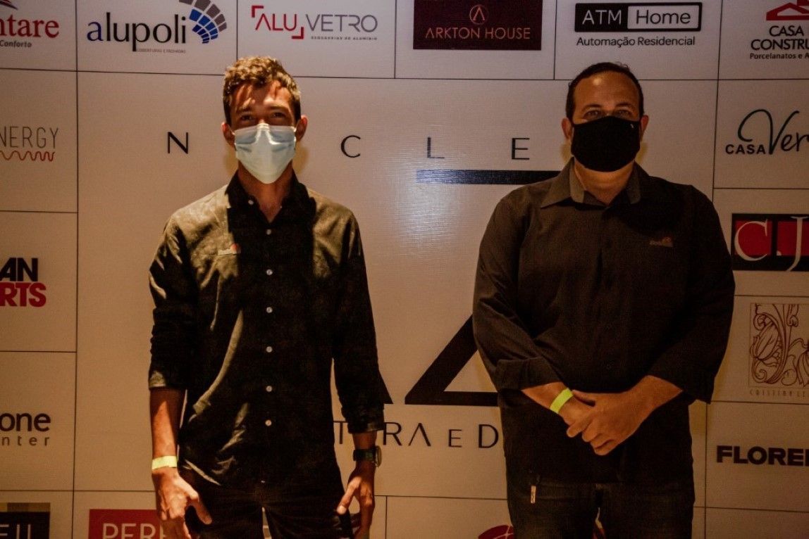 Dois homens usando máscaras faciais estão em frente a uma parede com logotipos de várias empresas