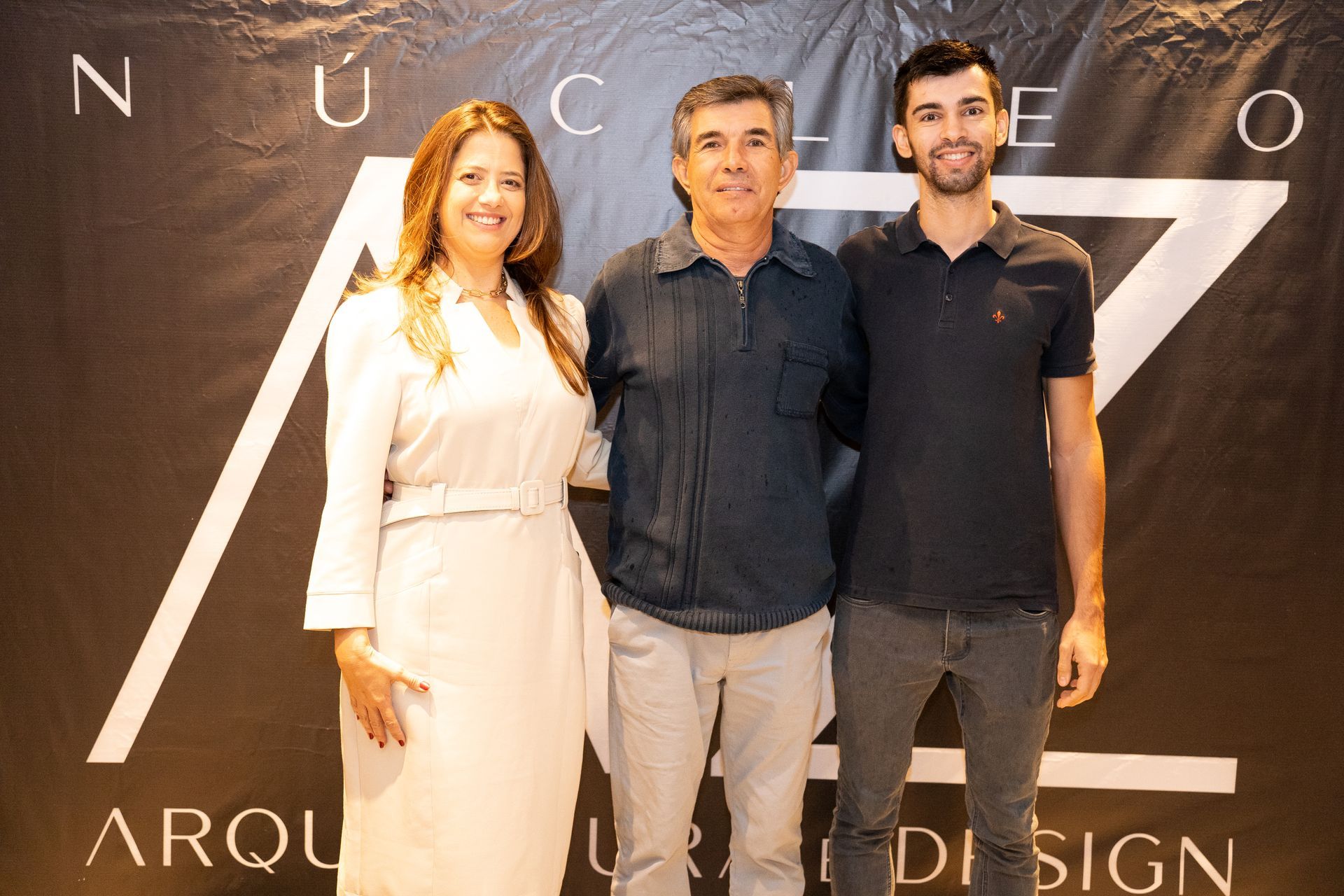 Três pessoas estão posando para uma foto em frente a uma placa que diz arqui design