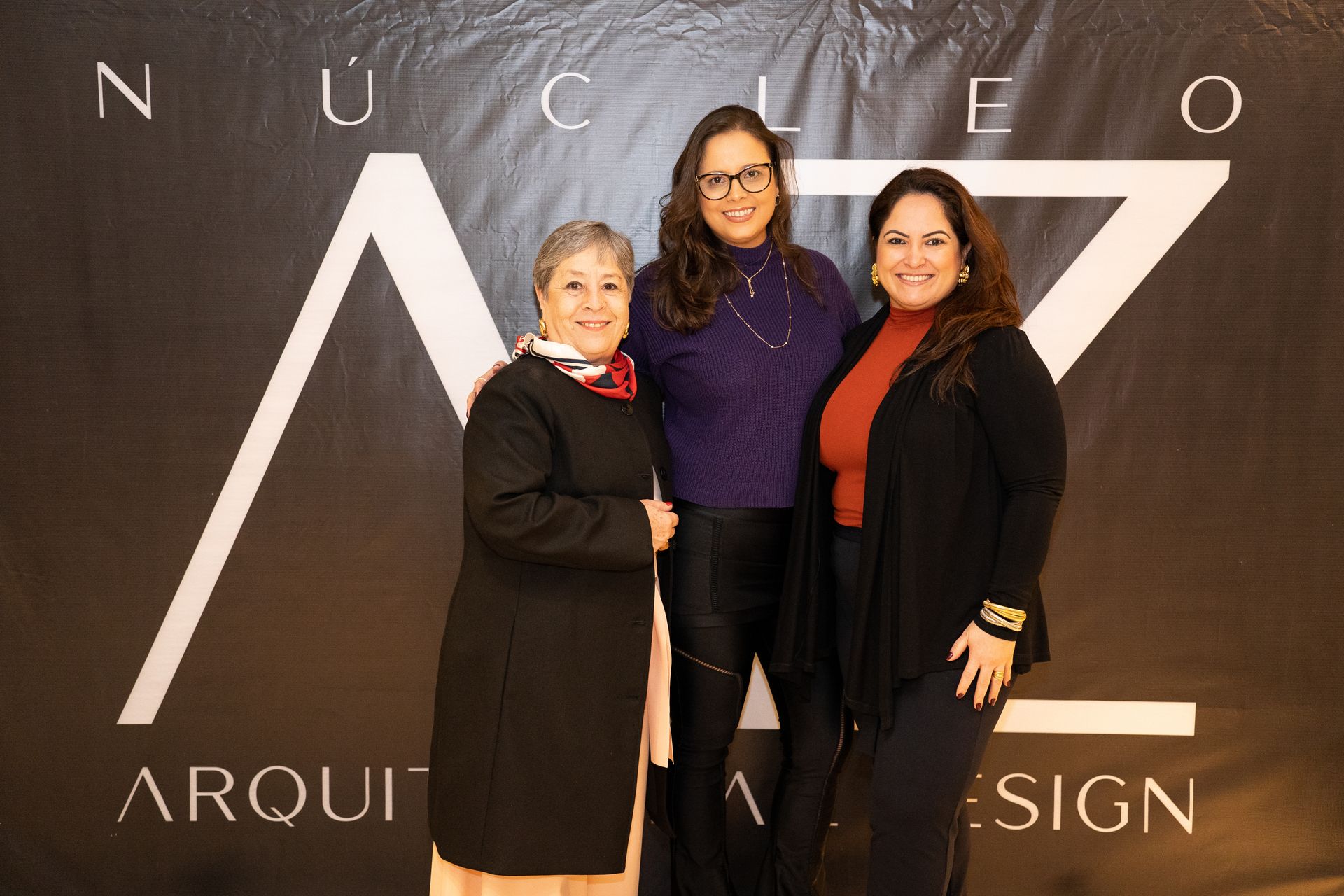 Três mulheres estão posando para uma foto em frente a uma placa que diz arqui design