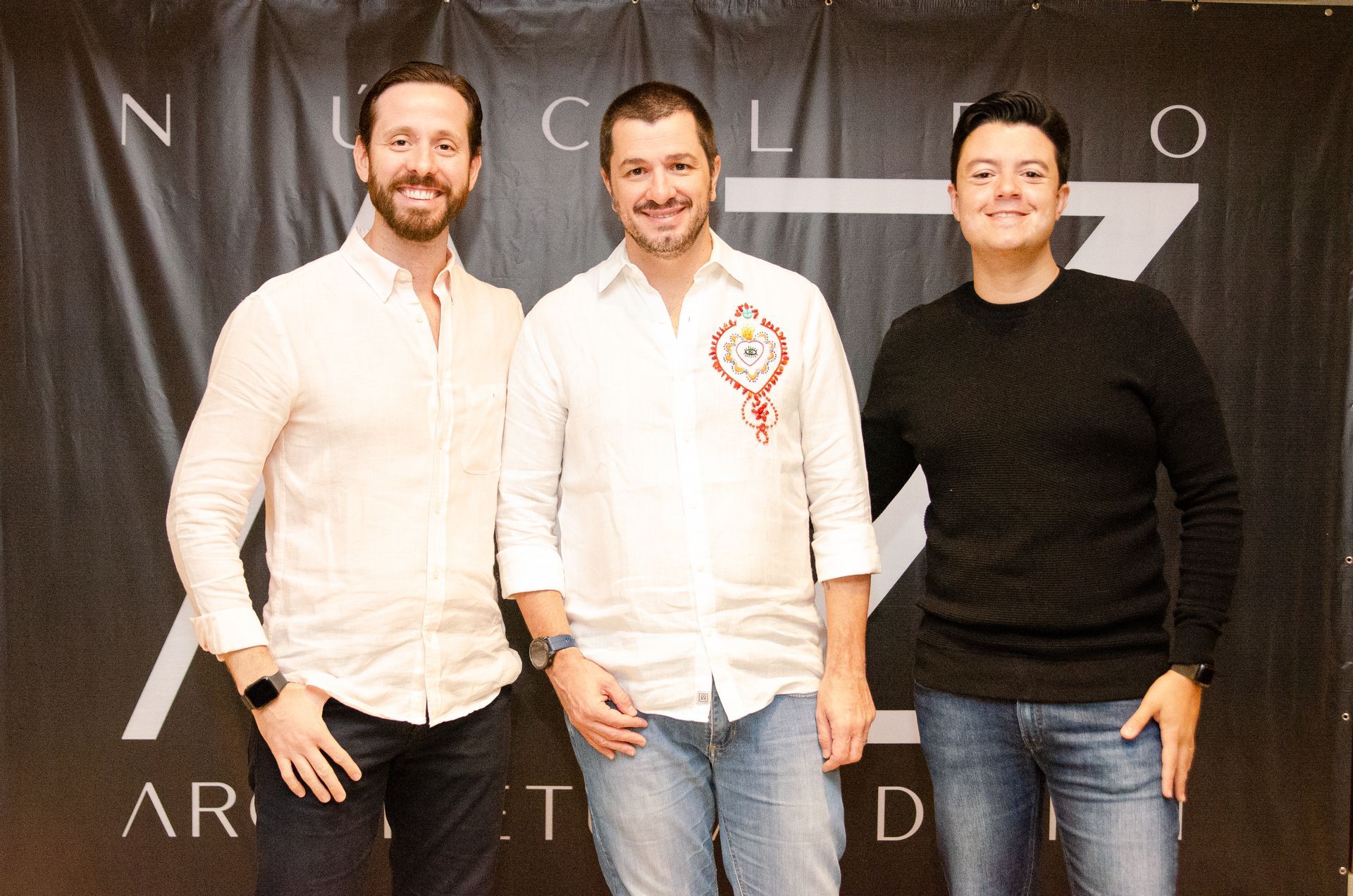 Três homens estão posando para uma foto em frente a uma faixa preta.