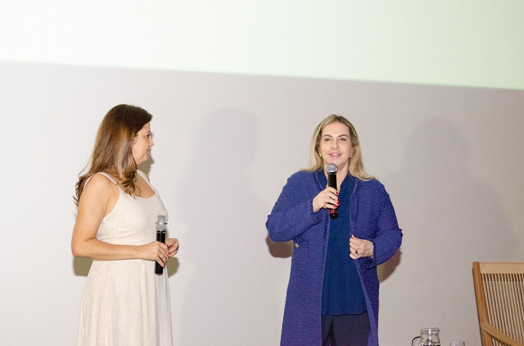 Duas mulheres estão de pé, uma ao lado da outra, em um palco, segurando microfones.