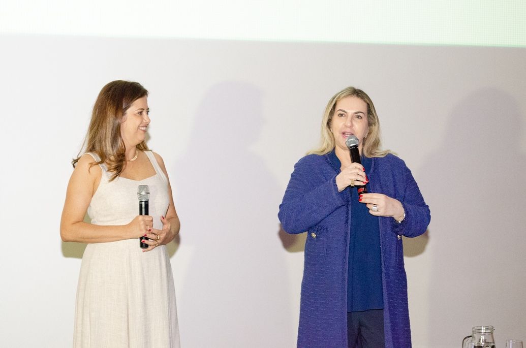 Duas mulheres estão de pé, uma ao lado da outra, segurando microfones.
