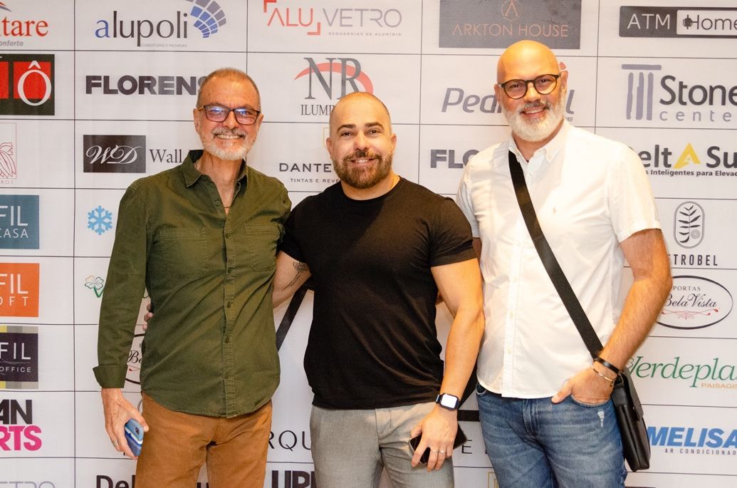 Três homens estão posando para uma foto em frente a uma parede com logotipos