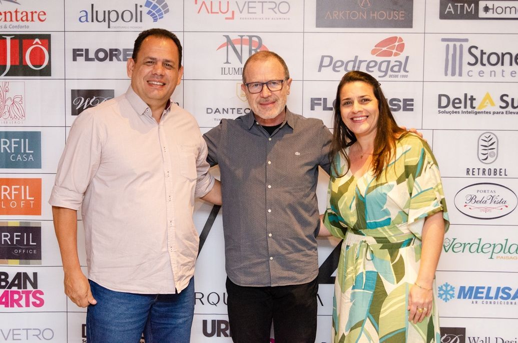 Três pessoas estão posando para uma foto em frente a uma parede com logotipos.