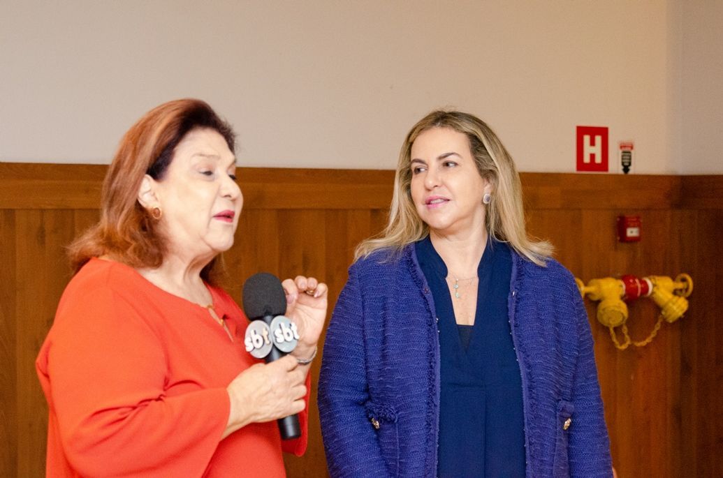 Duas mulheres estão de pé, uma ao lado da outra, falando em microfones.