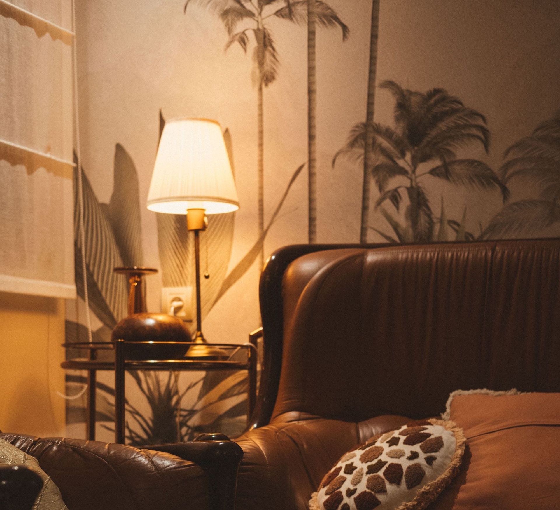 Cozy living room with leather sofa, lit table lamp, and palm-pattern wallpaper at dusk