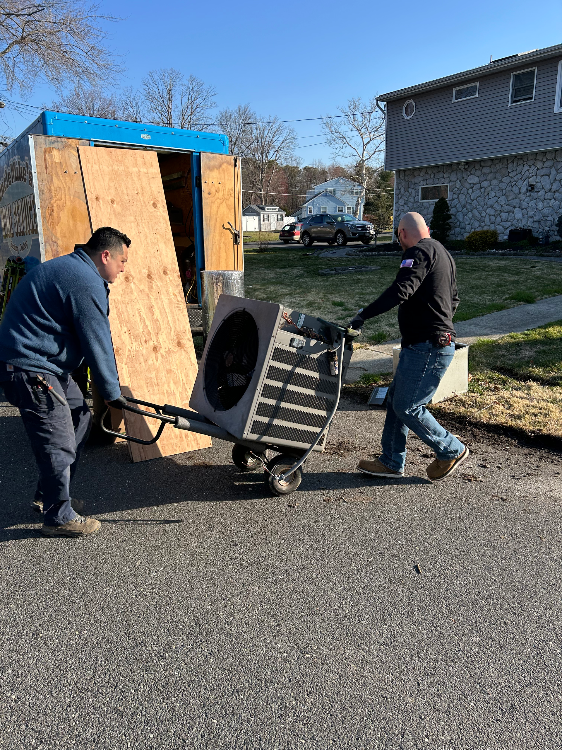 Two men wheel a large black barrel beside a wooden shed on a sunny residential street.