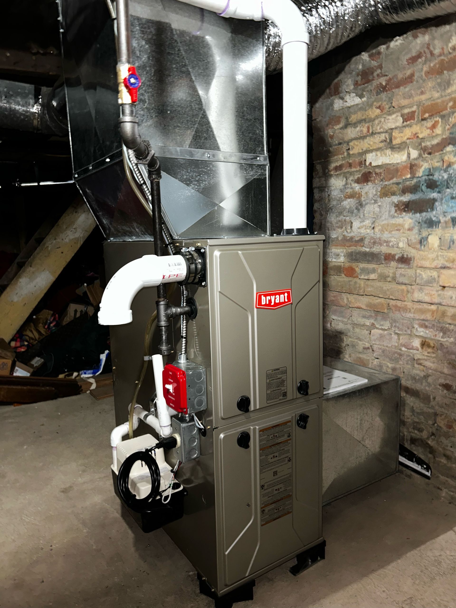Grey HVAC furnace with ductwork in a basement utility room beside a brick wall.