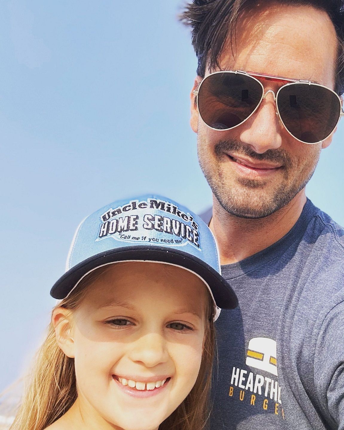 Smiling child in a blue cap and man in sunglasses taking a close selfie outdoors under a clear sky