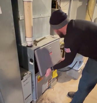 Man opening a gray furnace panel in a basement utility room
