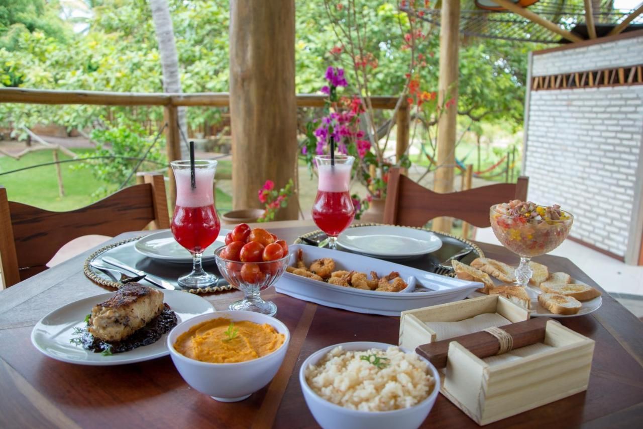 Uma mesa posta com uma refeição incluindo bebidas, peixe, acompanhamentos e petiscos em um deck ao ar livre com vista para a vegetação.