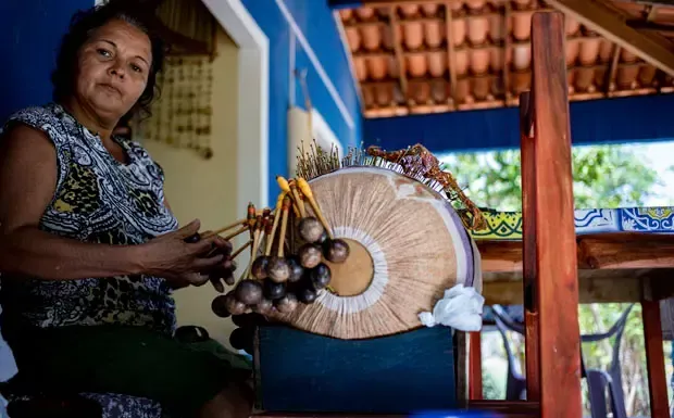 Uma artesã trabalha em uma delicada renda de bilros sobre uma almofada cilíndrica apoiada em um suporte de madeira em um ambiente externo e iluminado.