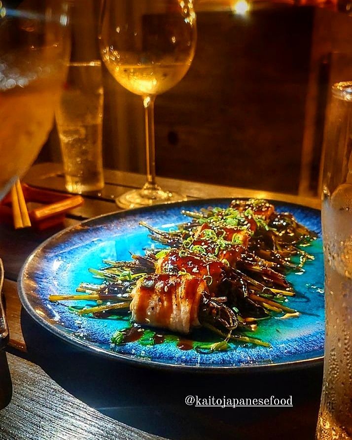 Um prato de legumes grelhados envoltos em carne, regados com molho, disposto sobre uma mesa escura com taças de vinho em um restaurante.
