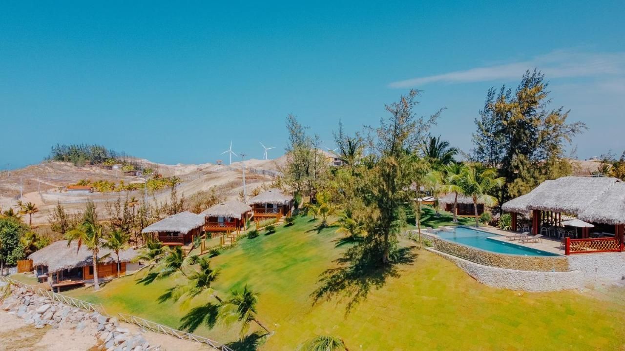 Um resort pitoresco com vilas de telhado de palha, piscina e árvores, tendo como pano de fundo dunas de areia e céu azul.
