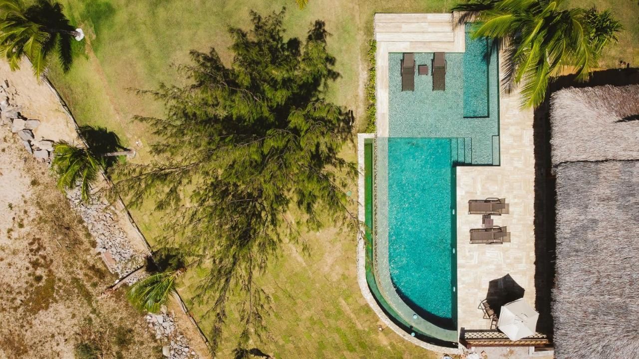 Vista aérea de uma piscina turquesa em um pátio ao lado de um gramado, árvores e um edifício com telhado de palha.