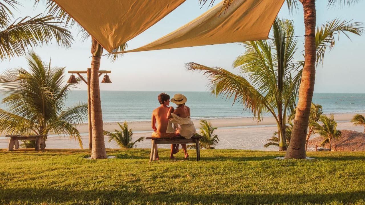 Um casal está sentado em um banco de madeira sob um toldo, com vista para uma praia ensolarada com palmeiras e águas calmas do oceano.