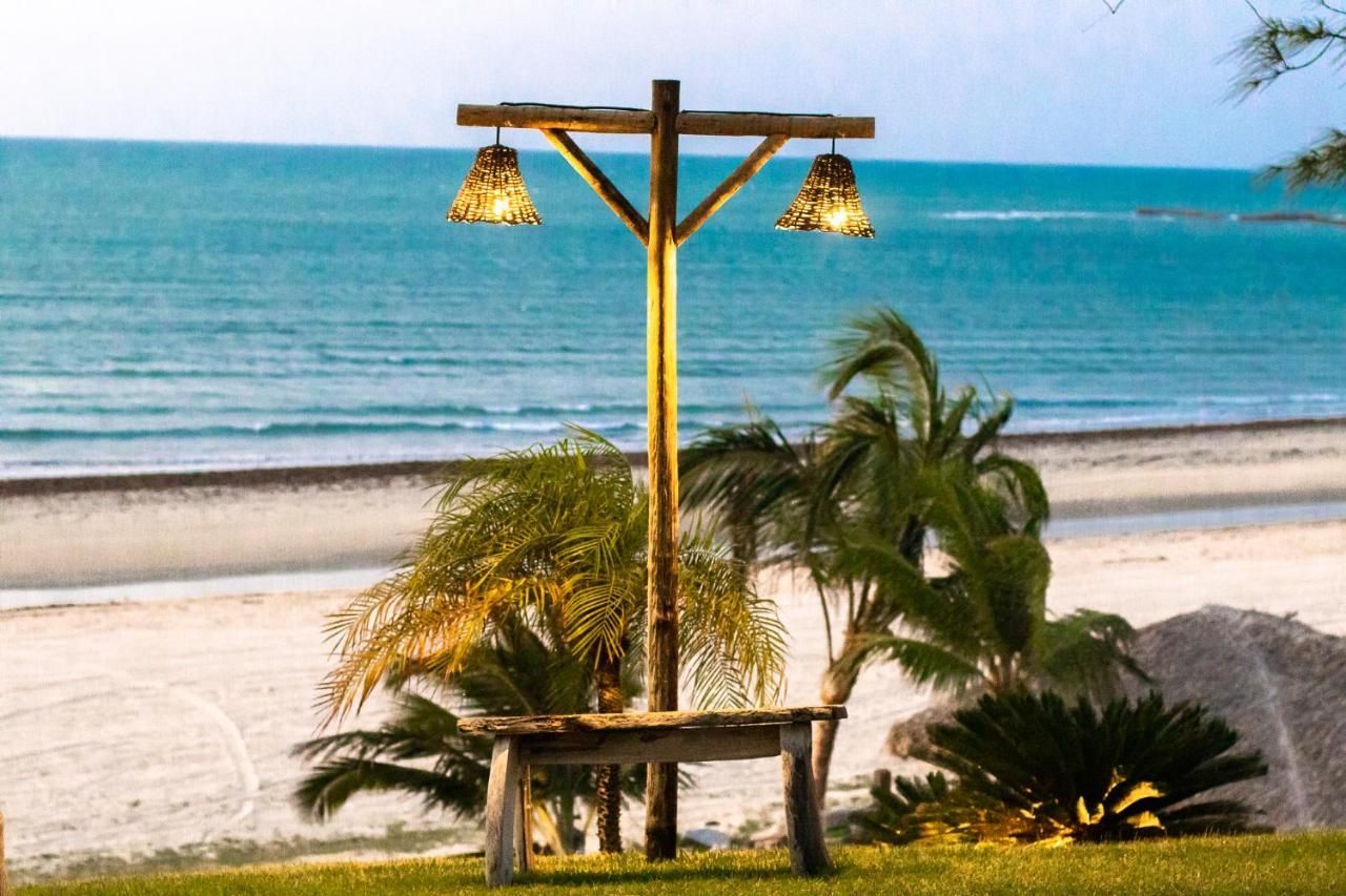 Um banco de madeira e um poste de luz com duas lanternas penduradas, com vista para uma praia de areia e o oceano ao entardecer.
