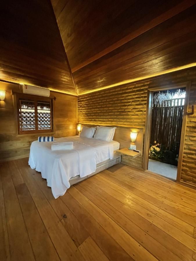 Um quarto de hotel aconchegante com paredes e piso de madeira, uma cama branca e uma porta que dá para uma área externa com iluminação suave.