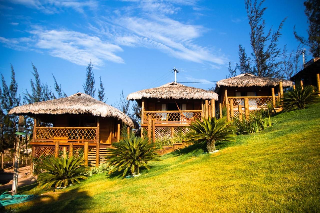 Três cabanas de madeira com telhados de palha estão situadas em uma encosta gramada e ensolarada, sob um céu azul claro.