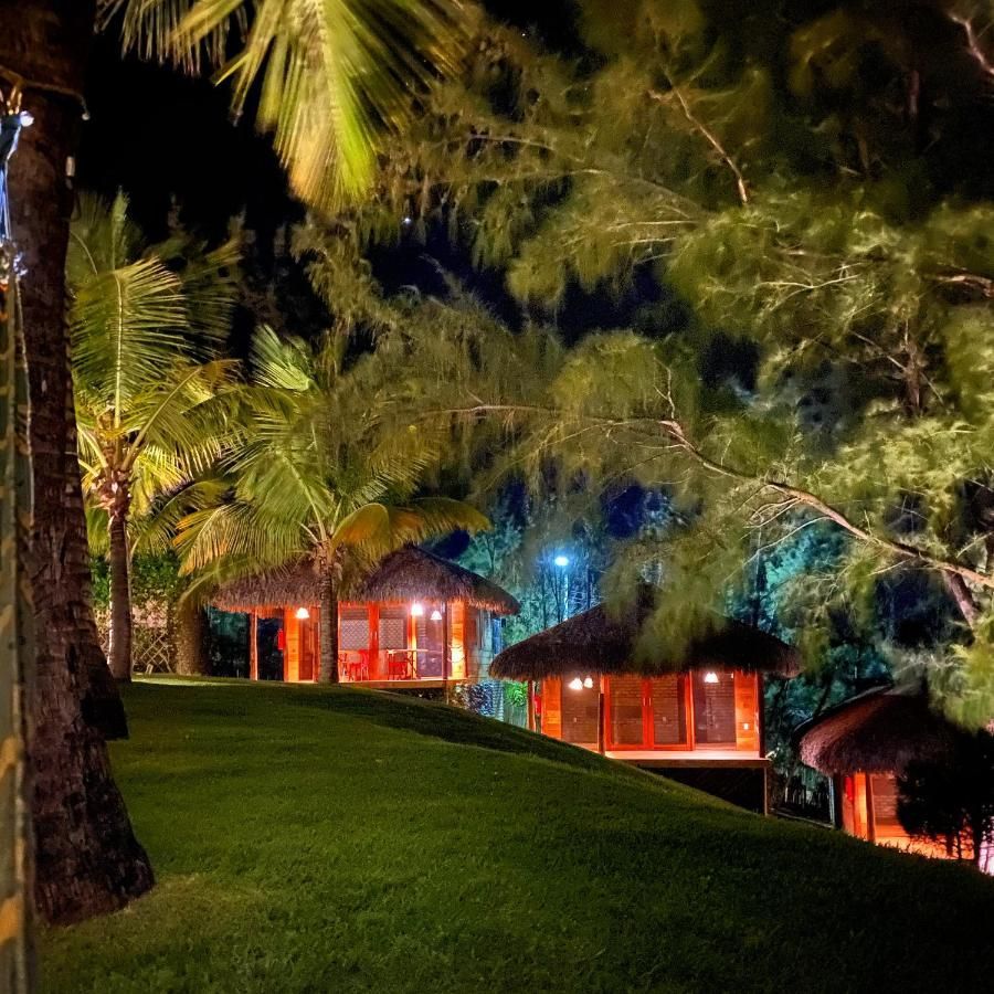 Pequenas cabanas de madeira iluminadas com telhados de palha, aninhadas à noite em uma encosta gramada, rodeadas por palmeiras.
