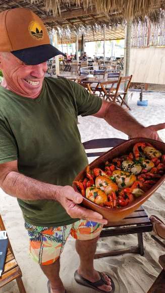 Uma pessoa sorridente, vestindo camisa e chapéu verdes, segura um prato de cerâmica com peixe e legumes cozidos em um café ao ar livre na praia.