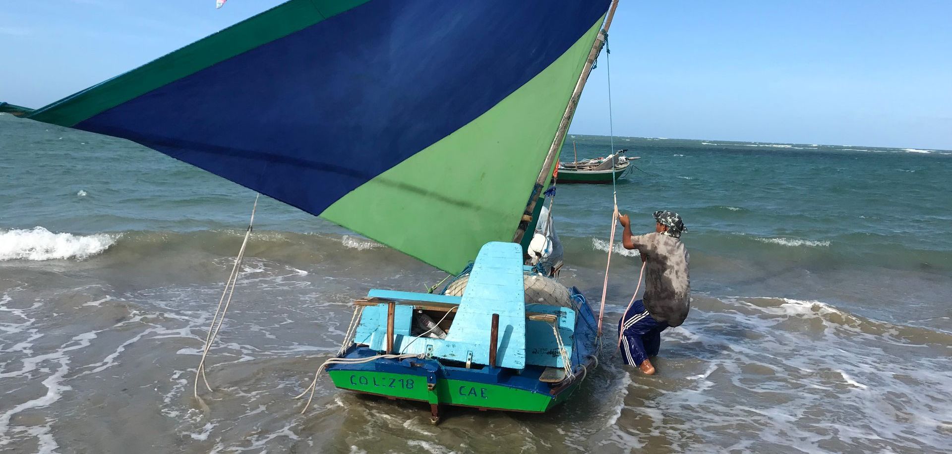 Em um dia ensolarado, uma pessoa empurra um pequeno veleiro com uma vela azul e verde para as ondas do oceano.