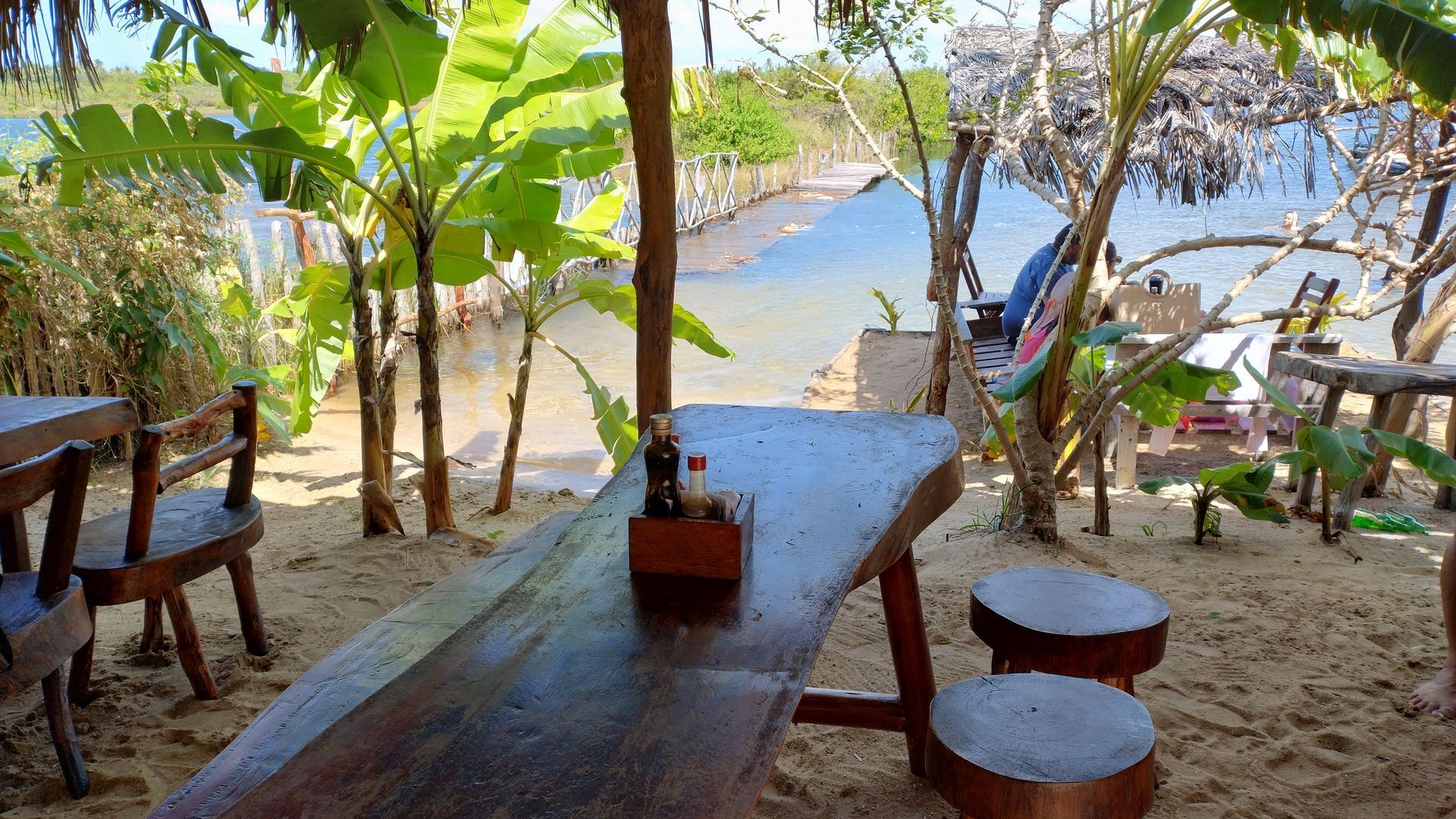 Uma área de jantar rústica ao ar livre, com mesas e bancos feitos de tocos de árvores, em uma praia de areia com vista para uma baía azul tranquila.